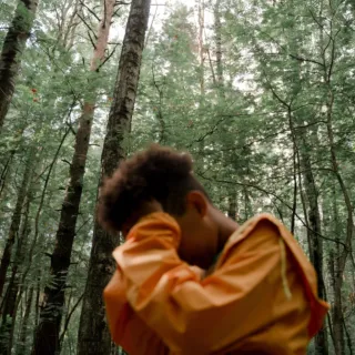 Person in orange jacket in a forest, covering face with arm. Tall trees in the background.