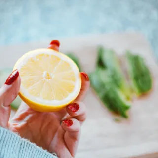 Hand holding half a lemon with red nails, avocado slices on a wooden board in the background.