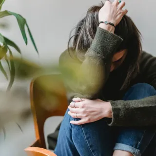 Person sitting on a chair, head down, wearing a green jumper and jeans, with plants nearby.