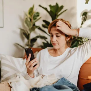 Woman in white top looking at phone, sitting on a sofa with plants in the background.