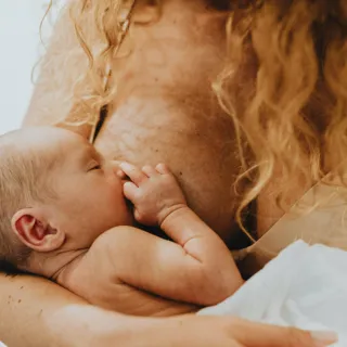 Baby breastfeeding, held by a person with curly hair, wrapped in a white cloth.