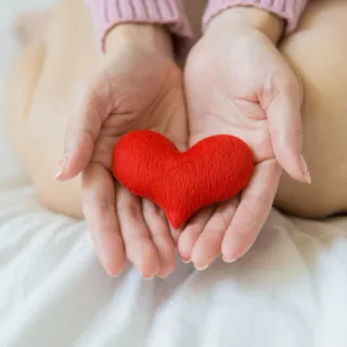 Hands holding a small red heart-shaped object on a white bed.