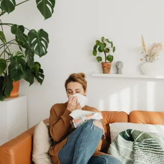 Woman sitting on a sofa, using a tissue. Indoor plants and decor on shelves in the background.