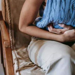 Person sitting on a sofa, holding a blue knitted hot water bottle to their stomach.