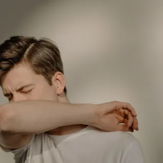 Person sneezing into their elbow, wearing a white shirt, against a neutral background.