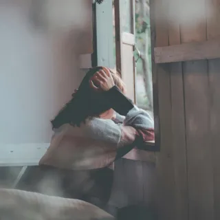 Person in a jumper looking out of a window, hand on head, indoors.