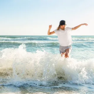 Person in white shirt and hat playing in ocean waves on a sunny day.