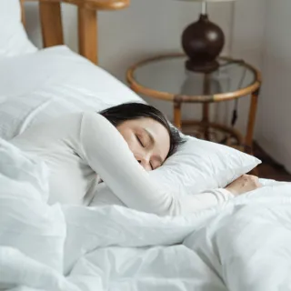 Person sleeping in a white bed next to a wooden chair and a bedside table with a lamp.