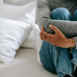 Person in grey jumper and jeans sitting on a bed, arms wrapped around knees.