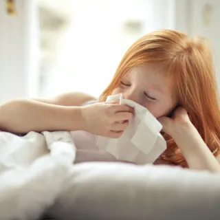 Child with red hair using a tissue in bed. Bright window in the background.