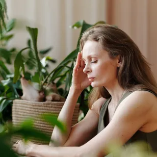 Woman sitting at a table, surrounded by green plants, resting her head on her hand.