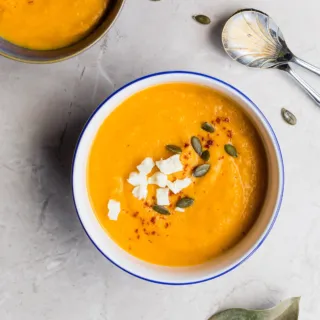 Bowl of orange soup with seeds and cheese on a marble surface, spoon and leaf nearby.