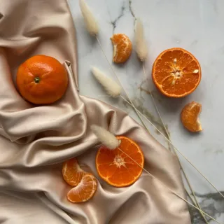 Whole and sliced tangerines on beige silk with dried grasses on marble surface.