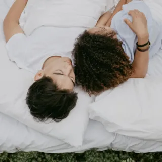 Two people lying on white bedding outdoors, surrounded by greenery.