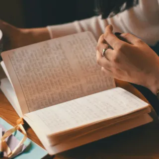 Person reading a notebook with handwritten text, holding a cup of tea on a wooden table.