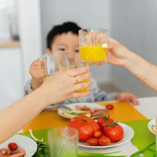 Three people clinking glasses of orange juice at a table with tomatoes and greens.