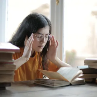 Person reading at a table with stacks of books, wearing glasses and an orange shirt.