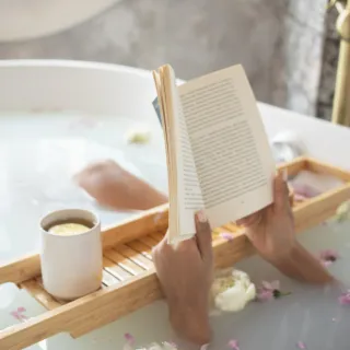 Person reading in a bath with flowers and a mug on a wooden tray.