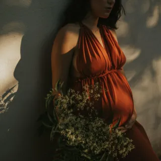 Pregnant woman in red dress holding flowers, standing against a sunlit wall.