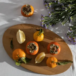 Persimmons, lemons, and mandarins on a wooden board with purple flowers on a white tablecloth.