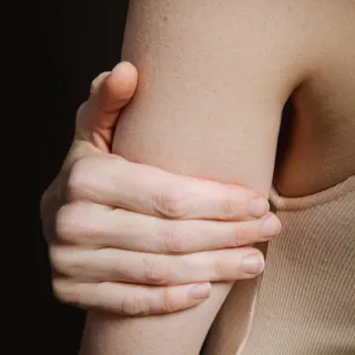 Person's hand holding their upper arm, wearing a beige top.