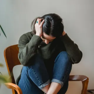 Person sitting on a chair, head down, wearing a green jumper and jeans.