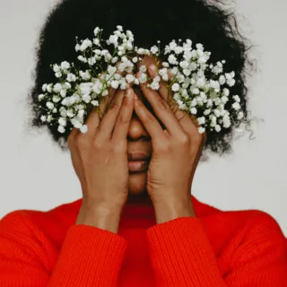Person in red jumper covering face with hands holding white flowers.