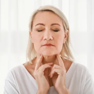 Woman with closed eyes touching her neck, standing in front of white curtains.