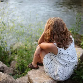 Person in a white dress sitting on rocks by a river, surrounded by greenery.