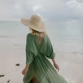 Woman in green dress and straw hat walking on a sandy beach by the sea.