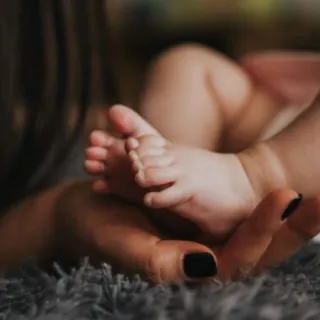 Baby's feet resting on a woman's hand with black nail polish, on a grey textured surface.