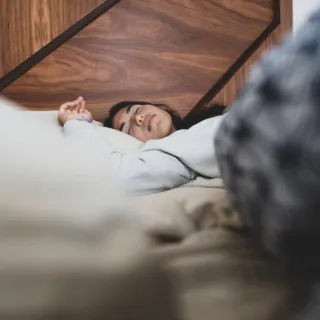 Person sleeping on a bed with a wooden headboard, wearing a light-coloured top.