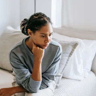 Person sitting on a bed in a bright room, looking thoughtful.