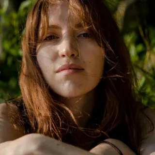 Woman with long hair sitting outdoors in sunlight, surrounded by greenery.