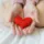 Hands holding a small red heart-shaped object on a white bed.
