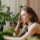 Woman sitting at a table, surrounded by green plants, resting her head on her hand.