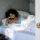 Woman in blue top sleeping in bed with white sheets, plant on bedside table.