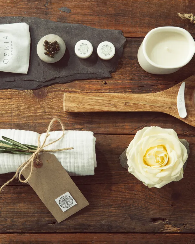Skincare items on wooden table: brush, cloth, candle, cream, rosemary, and a white rose.